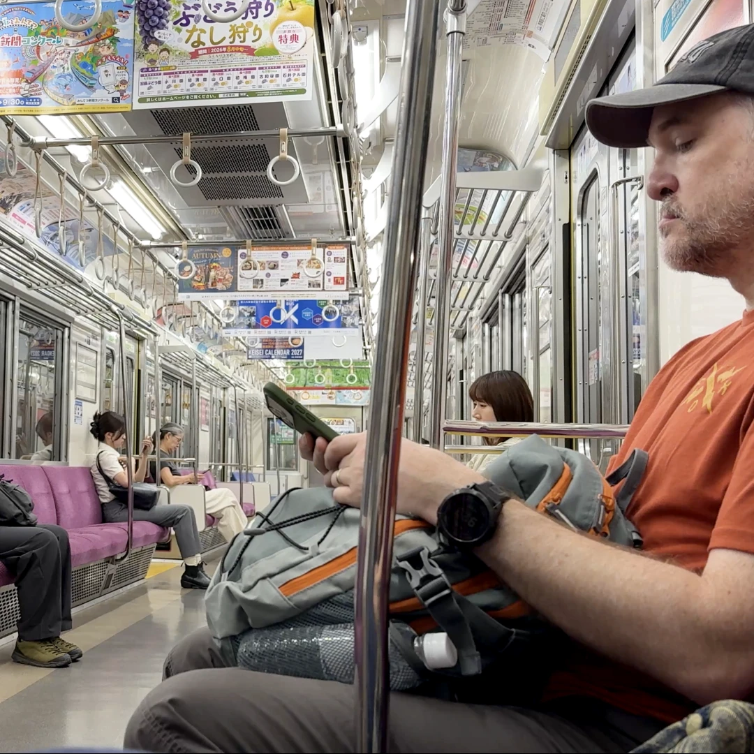 Passengers seated on a quiet Japanese commuter train with purple bench seats, hanging grab handles and colourful overhead adverts.