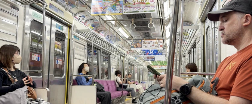 Passengers seated on a quiet Japanese commuter train with purple bench seats, hanging grab handles and colourful overhead adverts.