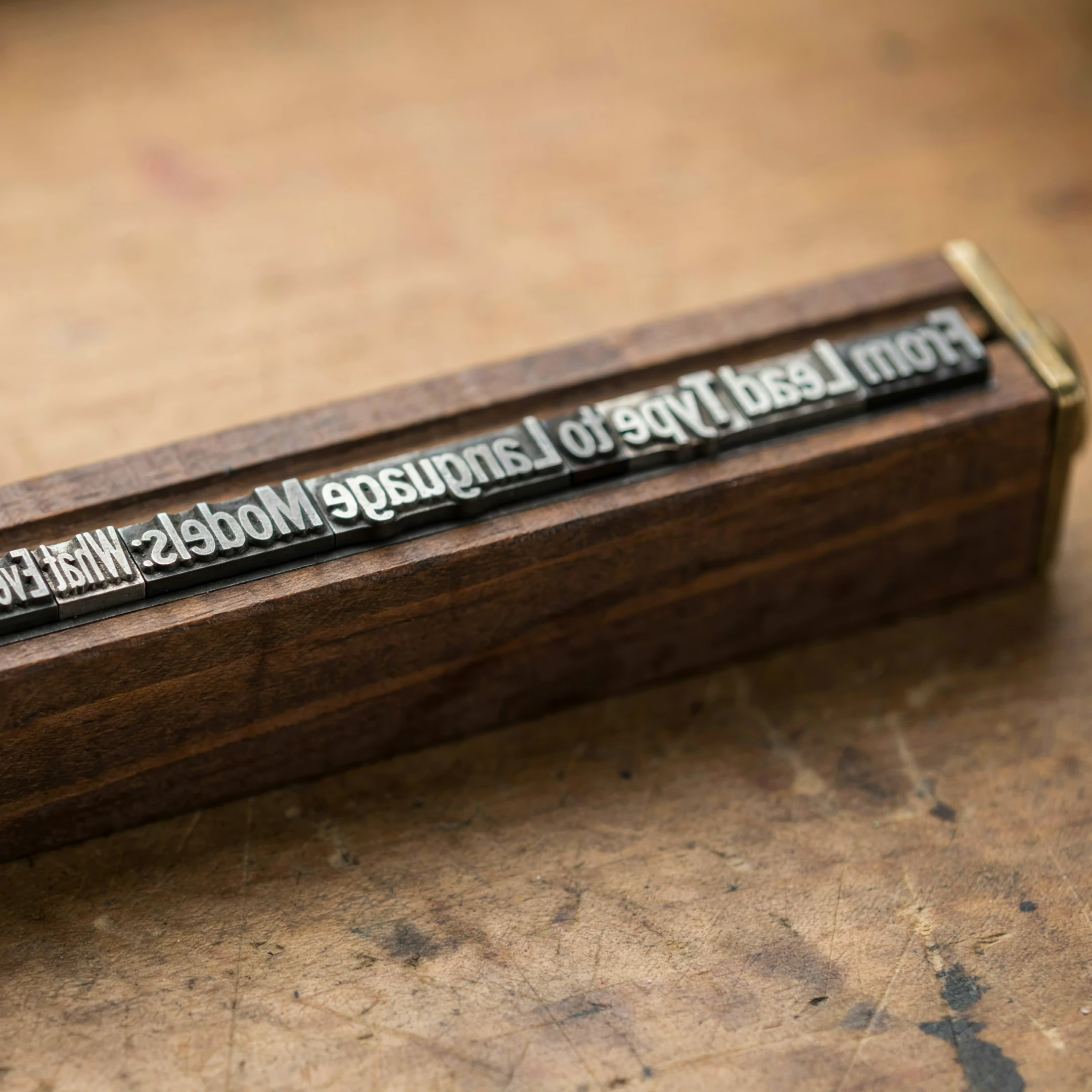 Mirror-image metal type in a wooden holder with brass ends on a worn, ink-stained workbench, spelling out the article title “From Lead Type to Language Models”